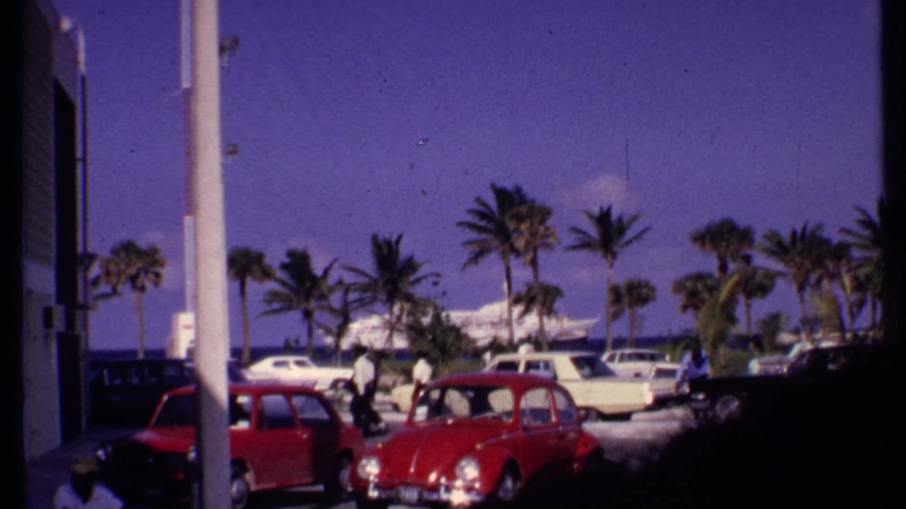 1969 Bahamas 8mm view from a coastal parking area with palm trees, classic cars, people, and a harbor ship in the distance.