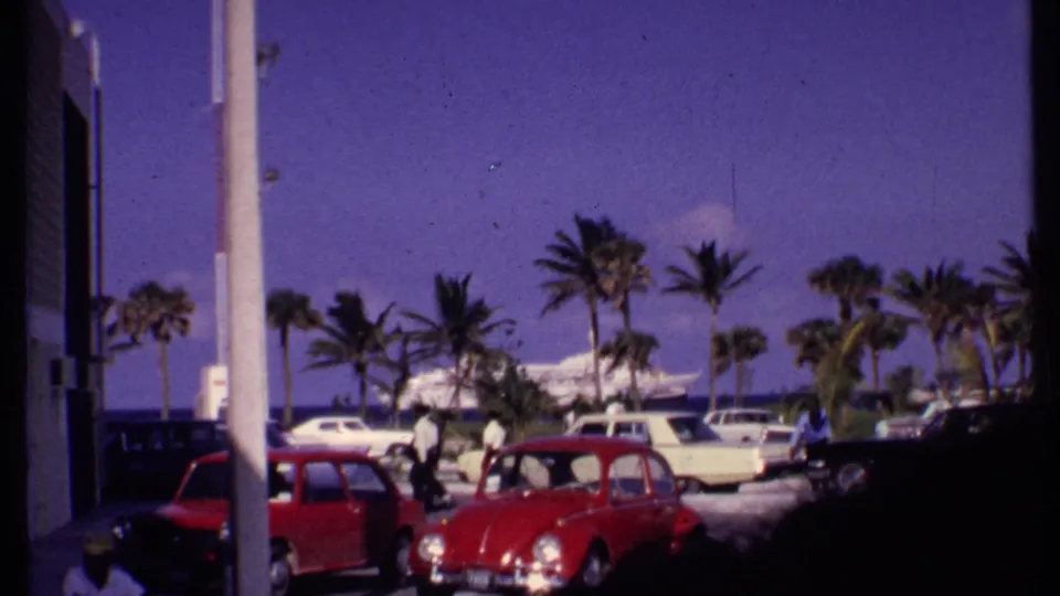 1969 Bahamas 8mm view from a coastal parking area with palm trees, classic cars, people, and a harbor ship in the distance.