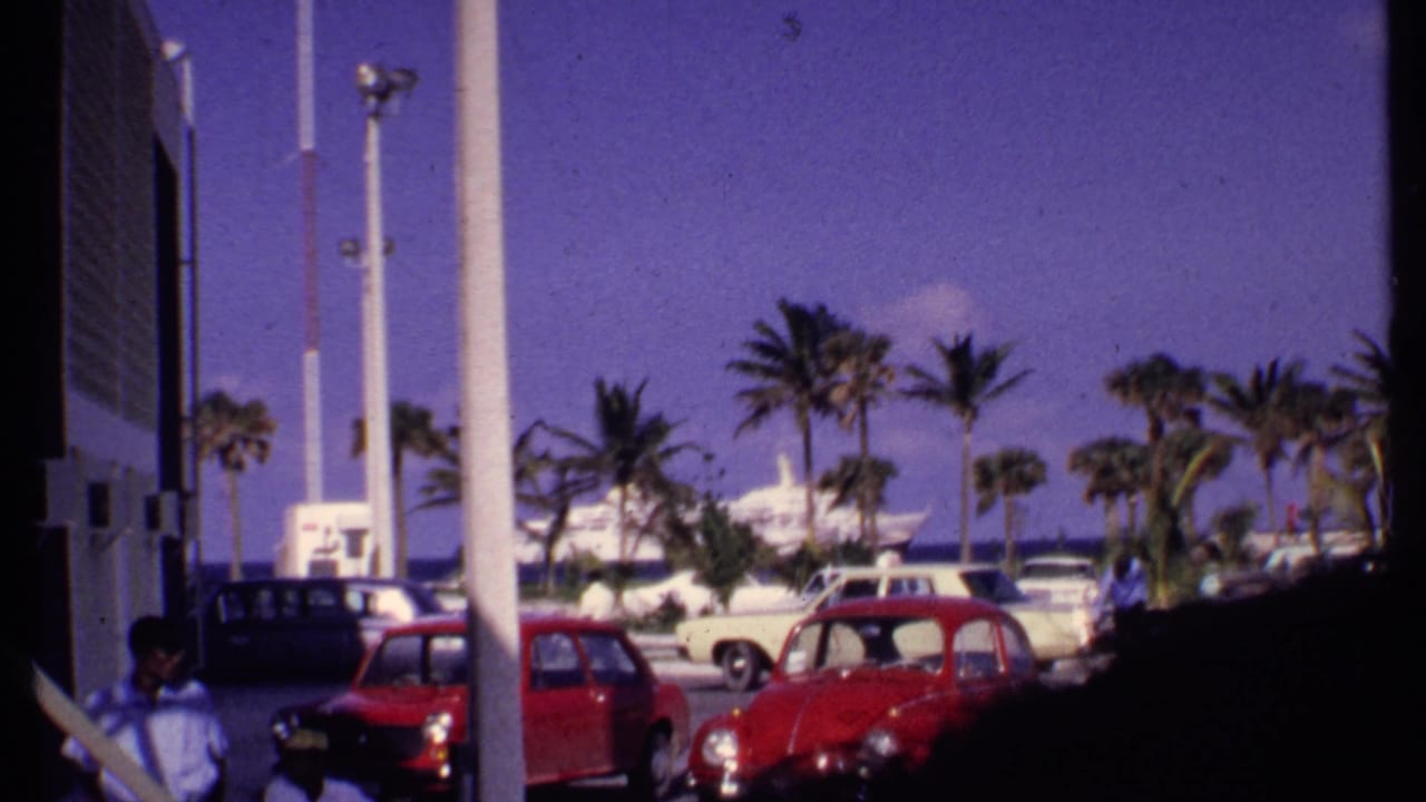 1969 Bahamas waterfront parking scene with a red Volkswagen-style Beetle, palms, distant people, and cruise ship activity near the harbor.