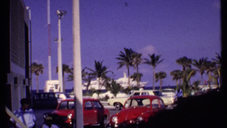 1969 Bahamas waterfront parking scene with a red Volkswagen-style Beetle, palms, distant people, and cruise ship activity near the harbor.