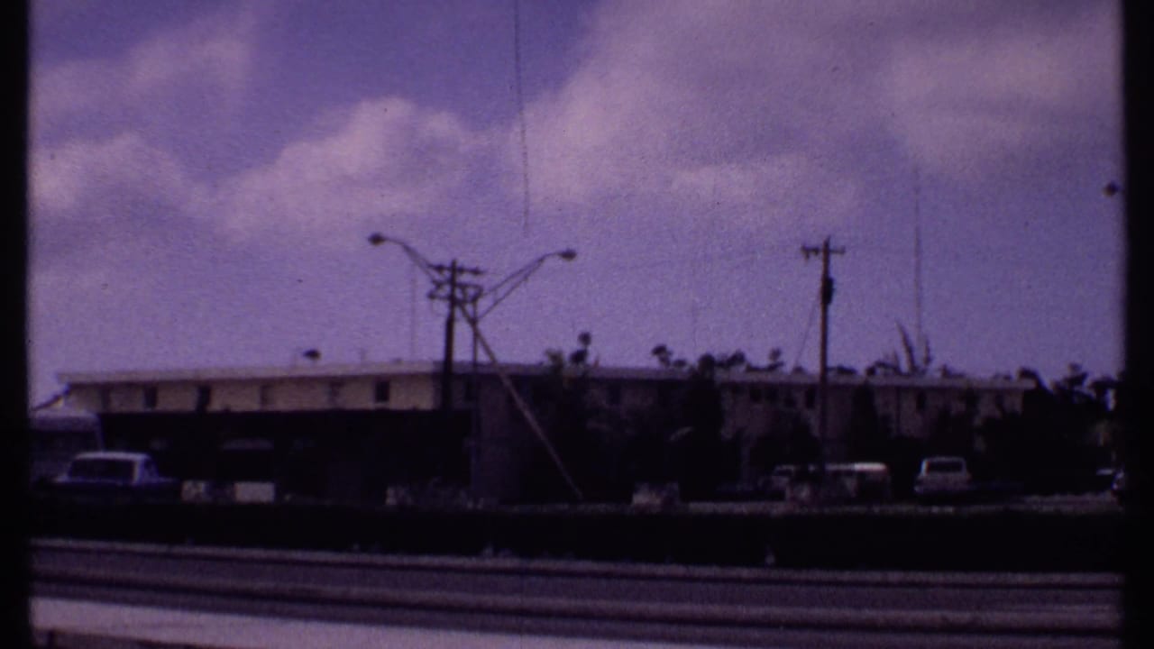 Roadside view with low buildings, parked cars, streetlights, utility poles, and cloudy sky.
