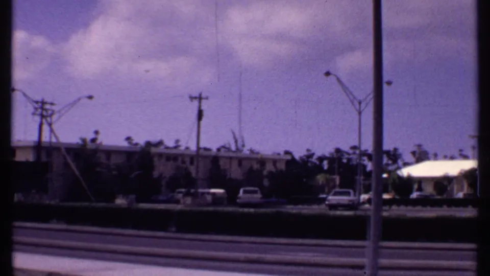 1969 Bahamas roadway view with low buildings, parked cars, streetlights, utility poles, and a distant communications tower.