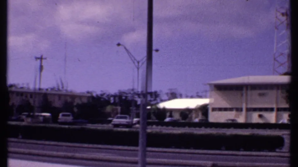 1969 Bahamas frame showing a road, parked cars, a white-roofed roadside building, streetlights, utility poles, and tower structure.