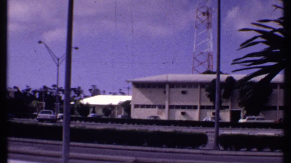 1969 Bahamas home-movie frame with a roadside building, parked car, tall streetlights, palm foliage, tower, and clouded sky.