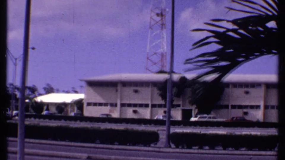 1969 Bahamas home-movie frame of a pale roadside building seen through palm fronds, with a hedge, light poles, and a tower in back.