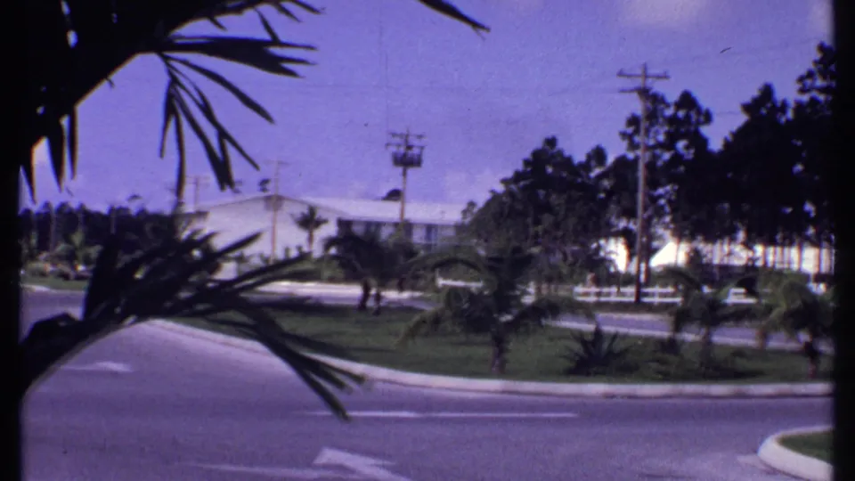 1969 Bahamas 8mm frame showing a curved street bordered by palms, lawns, utility poles, wires, trees, and a white low-rise building.