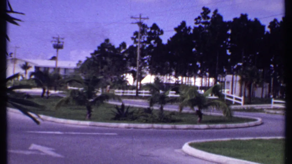 1969 Bahamas roadside view with a curved paved road, white fence, small palms, utility poles, trees, and low white buildings under a bright sky.