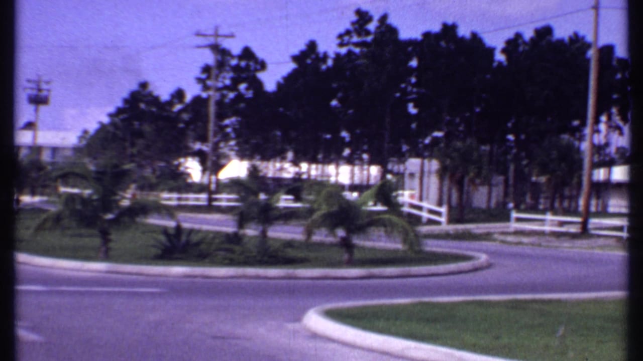 1969 Bahamas home-movie frame showing a curved roadside, white rail fence, palms, low bungalows, and dense trees.