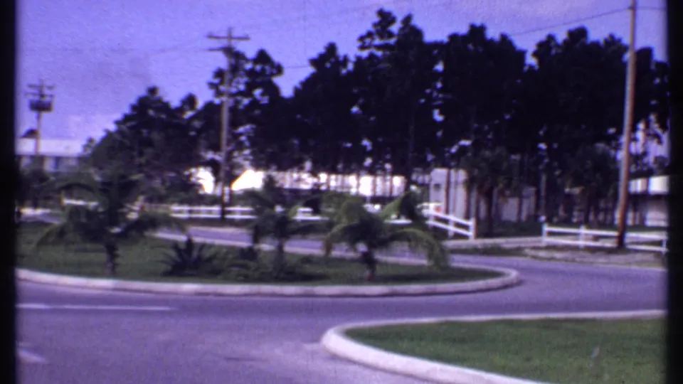 1969 Bahamas home-movie frame showing a curved roadside, white rail fence, palms, low bungalows, and dense trees.