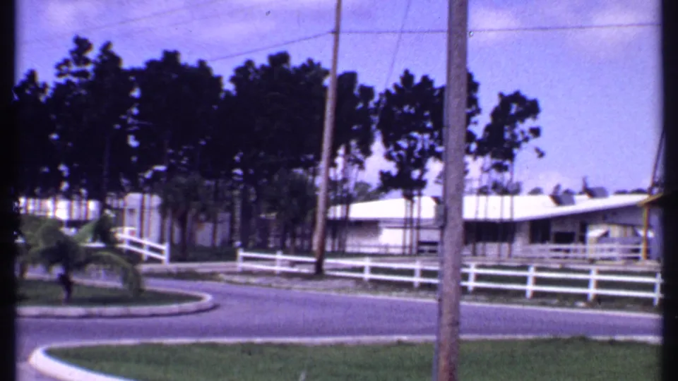 1969 Bahamas frame with white fencing, utility poles, small palms, low white-roofed bungalows, and a tree line.