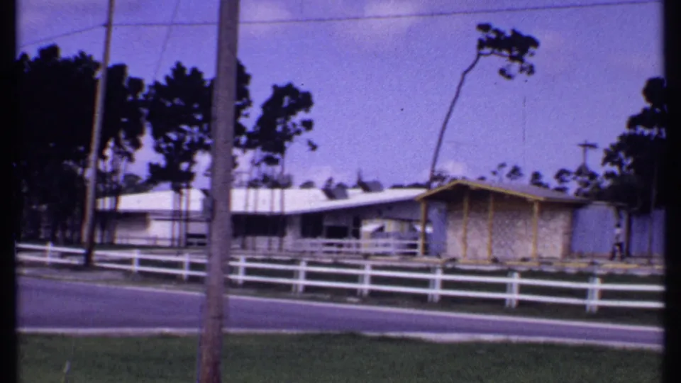 1969 Bahamas frame showing a roadside fence, utility pole, yellow-trimmed bungalow, and one distant pedestrian.