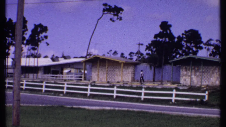 1969 Bahamas home-movie frame of a yellow-trimmed bungalow, white fence, roadside grass, trees, and a distant walker.
