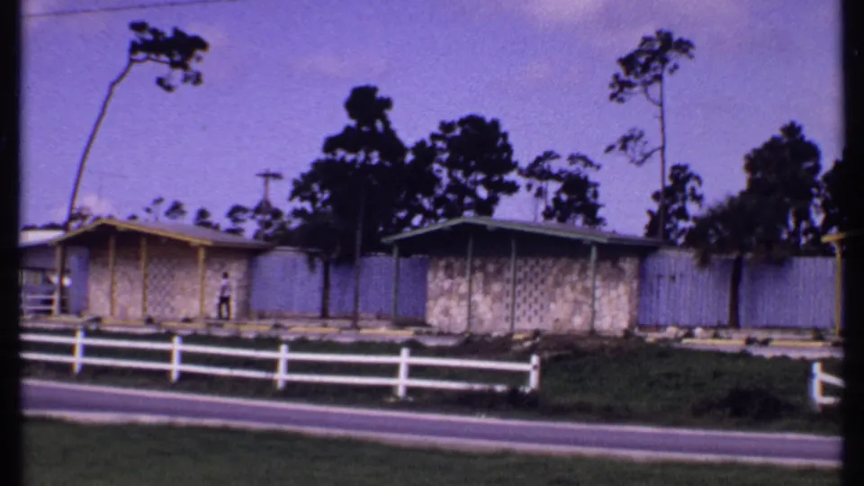 1969 Bahamas frame with modest single-story houses, a white roadside fence, trees, grass, and a small person near the building wall.