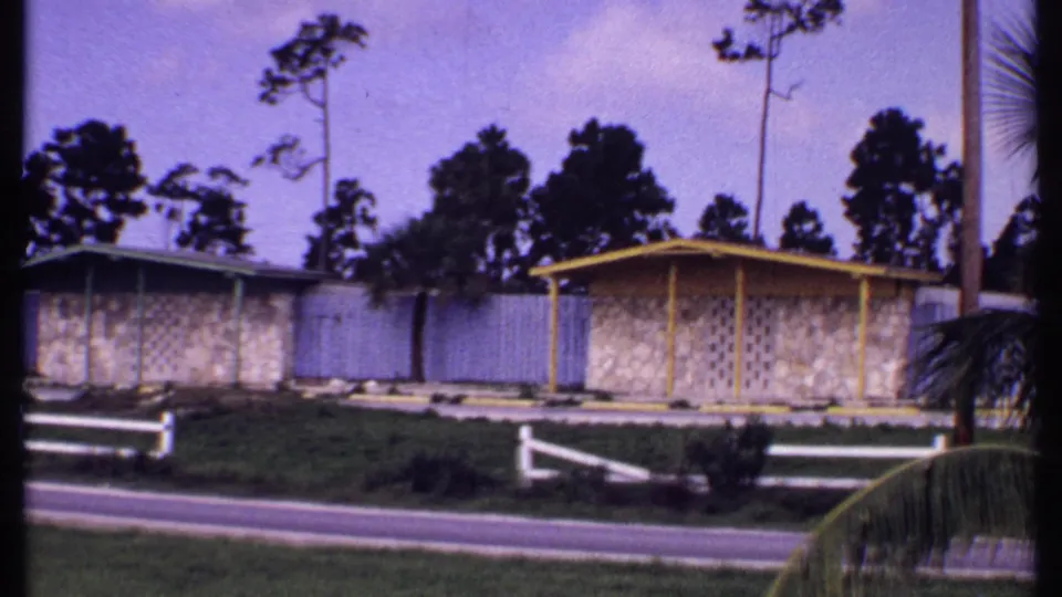 1969 Bahamas frame showing a low house with yellow trim, white fence, paved roadside, trees, grass, and palm fronds at the edge.