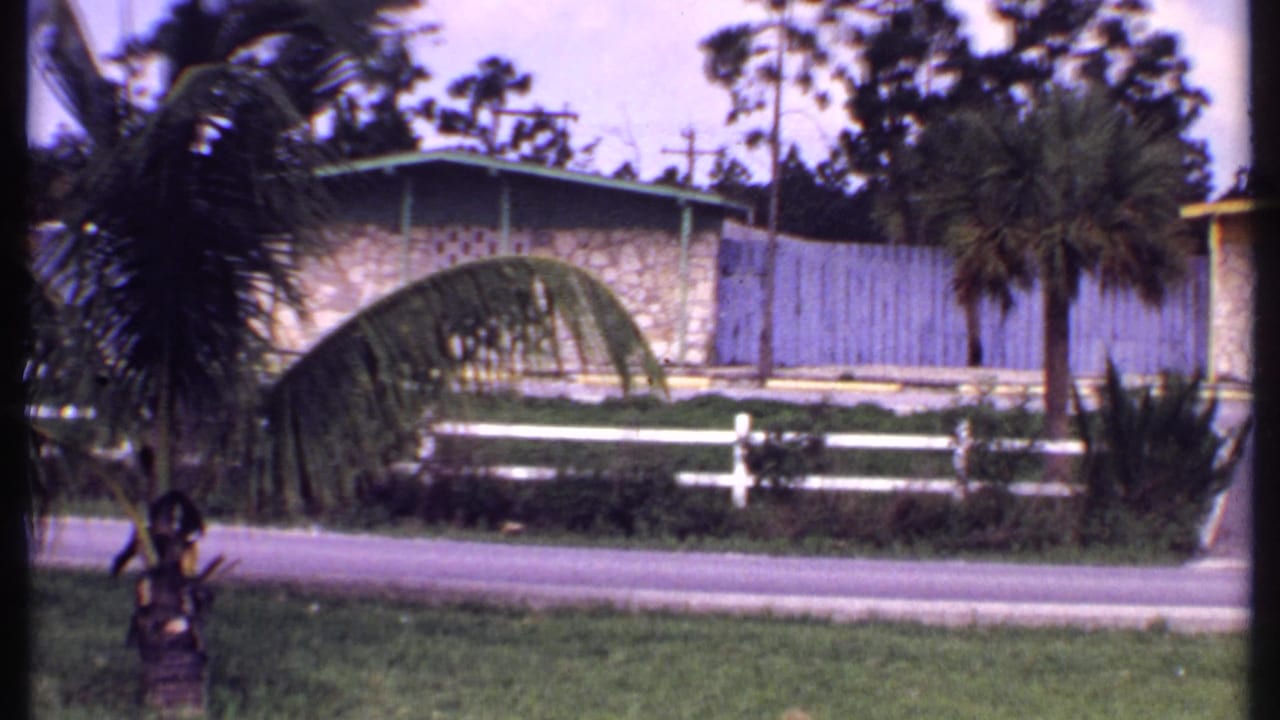 1969 Bahamas 8mm frame showing a tan stone bungalow with green trim, palms, a white fence, lawn, and roadside foreground.