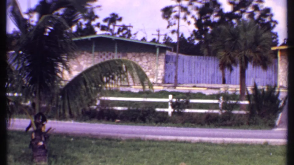 1969 Bahamas 8mm frame showing a tan stone bungalow with green trim, palms, a white fence, lawn, and roadside foreground.