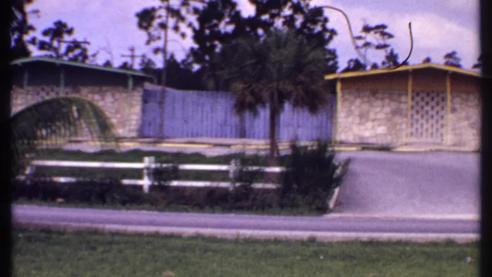 1969 Bahamas 8mm frame with a tan bungalow, driveway, palm trees, white fence sections, and landscaped roadside greenery.