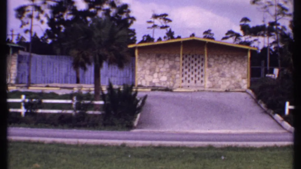 1969 Bahamas 8mm frame showing a tan bungalow with yellow trim, paved driveway, palms, blue fence, white fence, and lawn.