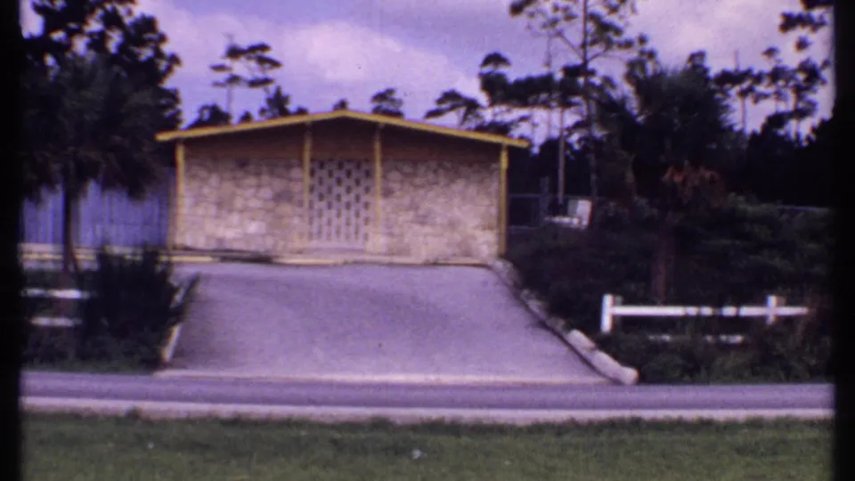 1969 Bahamas 8mm frame of a tan bungalow beyond a driveway, with palm trees, hedges, white fence, road, and bright sky.