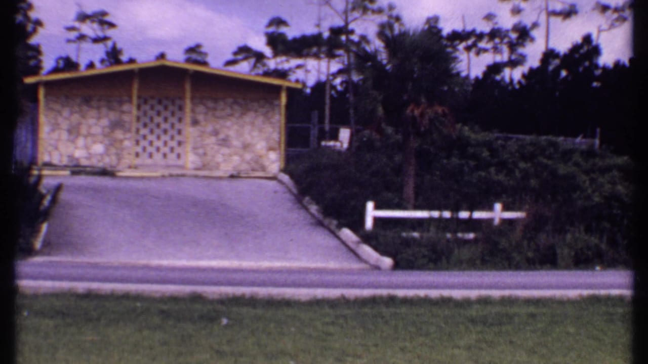 Bahamas 1969 home-movie frame showing a low stone-front roadside building, palms, hedges, pavement, and soft 8mm color fading.