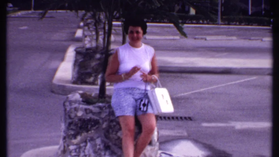 Bahamas 1969 frame of a woman seated outdoors near a stone planter, holding a white handbag on a paved tropical street.
