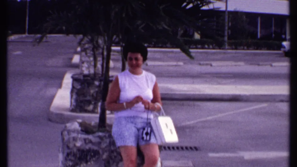Bahamas 1969 home-movie frame showing the woman seated by a stone planter, with parking lines, pavement, and roadside buildings behind her.