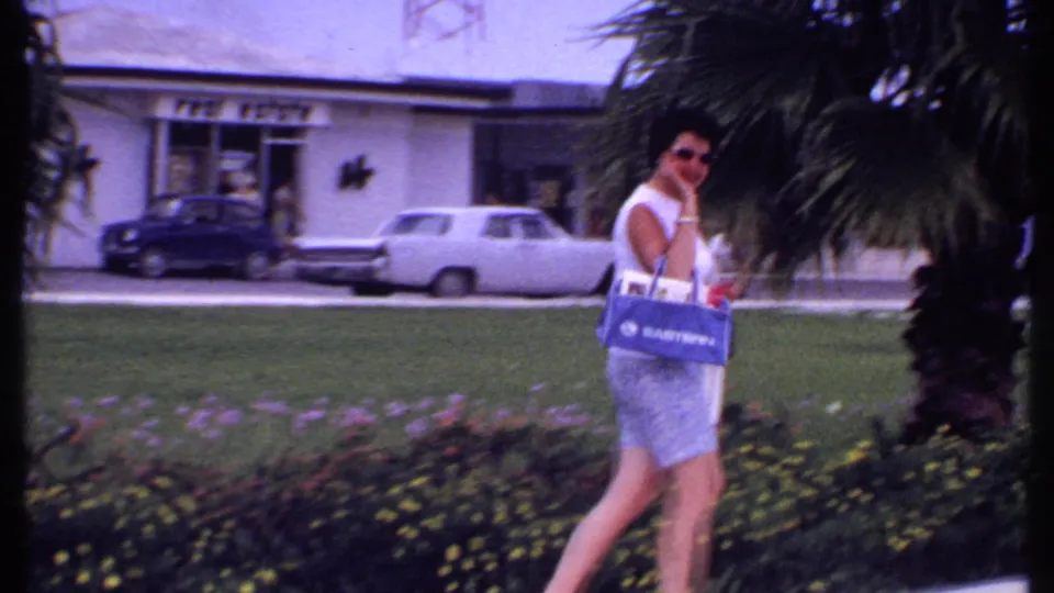 A woman in a white sleeveless top and patterned shorts carries shopping bags through a 1969 Bahamas tropical shopping-center garden.