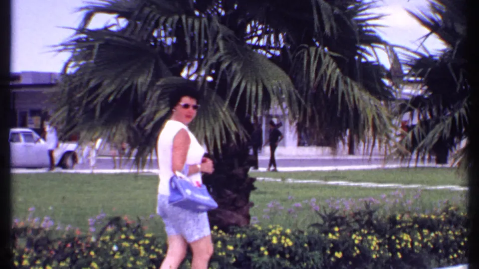 A woman holding a blue handbag pauses near a palm tree in the 1969 Bahamas shopping-center scene, with lawn and storefronts behind her.