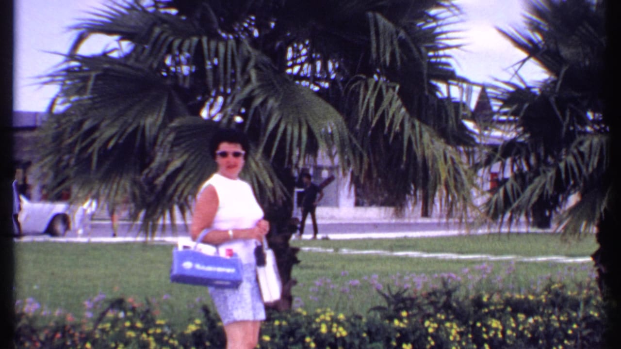 In 1969 Bahamas home-movie footage, a woman in sunglasses carries bags near palm trees, flowers, lawn, parked cars, and a low commercial building.