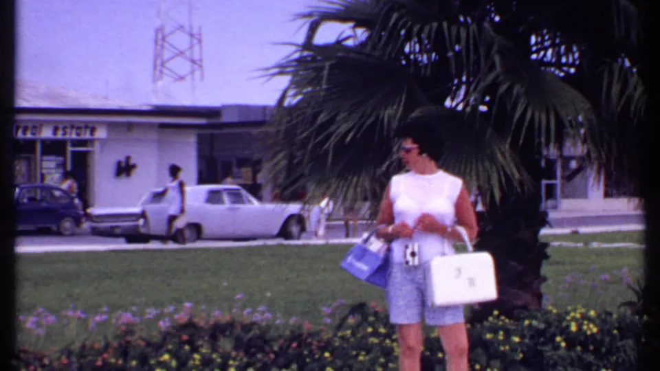 In the Bahamas in 1969, a woman in sunglasses walks near a palm tree and lawn with a white car and building signage visible behind her.