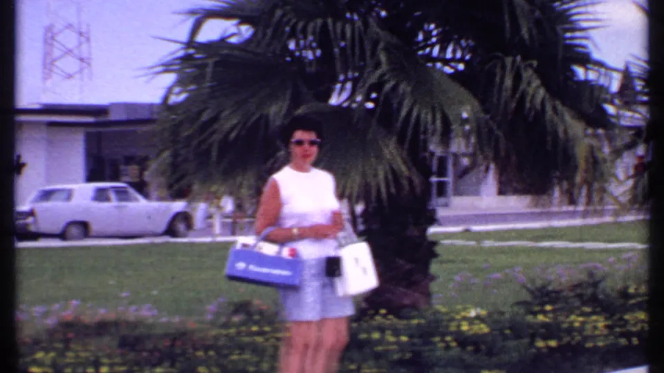 A 1969 Bahamas frame shows a woman in sunglasses holding shopping bags beside palm fronds, flower beds, grass, and a parked white car.