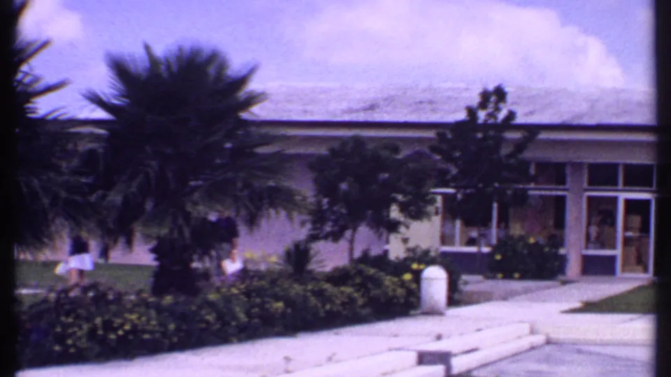 1969 Bahamas frame showing a low public-facing building, broad windows, palms, flower beds, steps, and distant pedestrians.