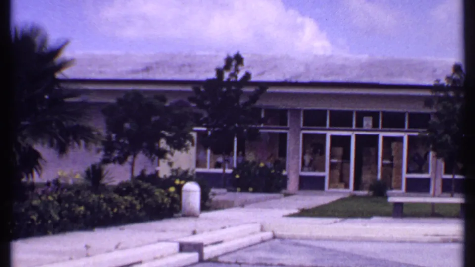 1969 Bahamas home-movie frame with a low glass-fronted building, tropical landscaping, steps, walkway, and bench.
