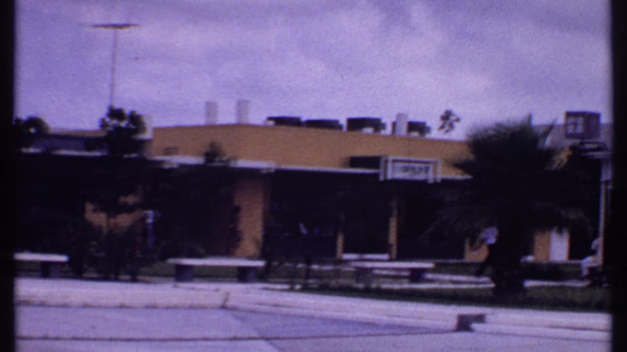 1969 Bahamas 8mm frame showing a low outdoor shopping center with palms, benches, pedestrians, and a visible storefront sign.