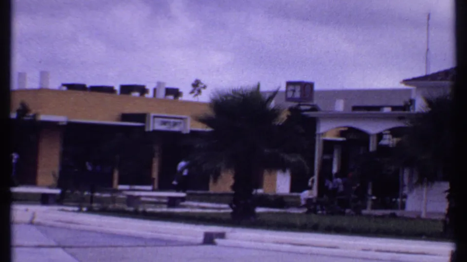 1969 Bahamas home-movie view of a shopping-center frontage with palms, benches, pedestrians, signage, and an arched entry area.
