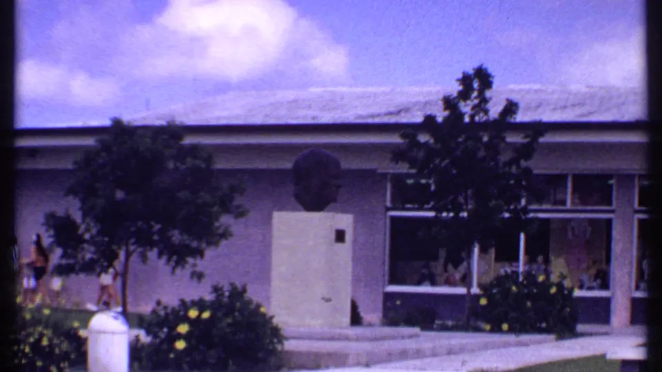 1969 Bahamas 8mm frame showing a low building, a bust on a pedestal, landscaped shrubs, small trees, and pedestrians nearby.