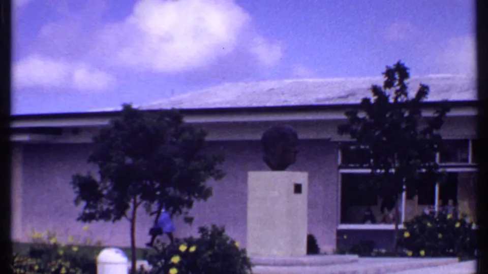 1969 Bahamas home-movie frame of a building exterior with a pedestal bust, trees, tropical shrubs, walkway, and a passerby.