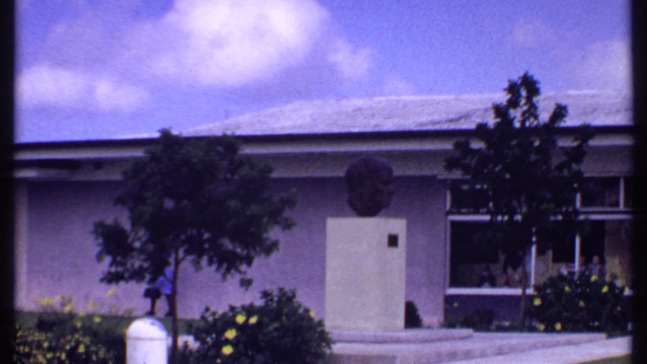 1969 Bahamas home-movie frame showing people walking outside a low retail building with lawn, trees, and broad windows.