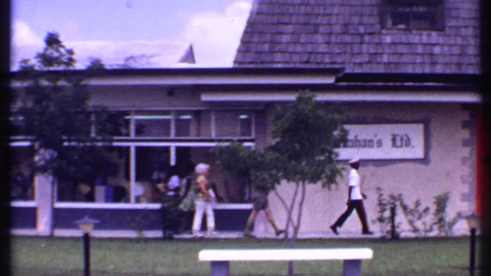1969 Bahamas home-movie view of pedestrians walking outside Kahan's Ltd., with windows, lawn, and a bench in front.
