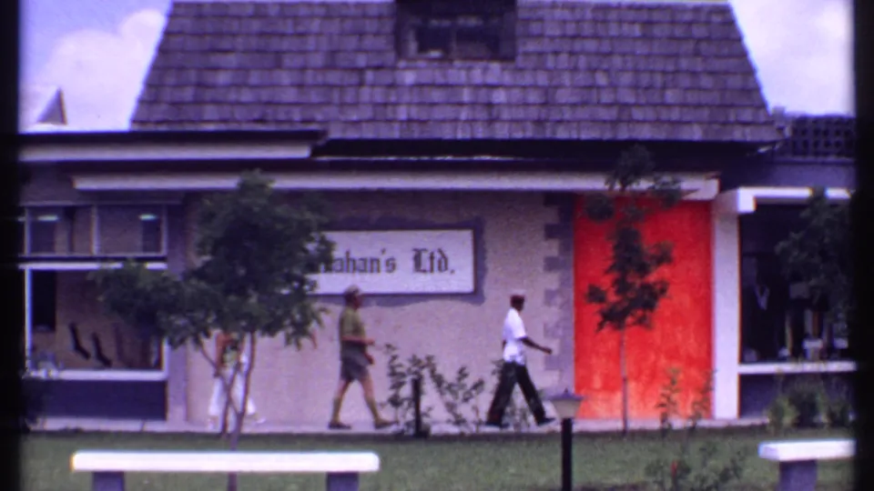 1969 Bahamas frame showing pedestrians outside the Kahan's Ltd. building beside a red wall panel and small landscaped area.