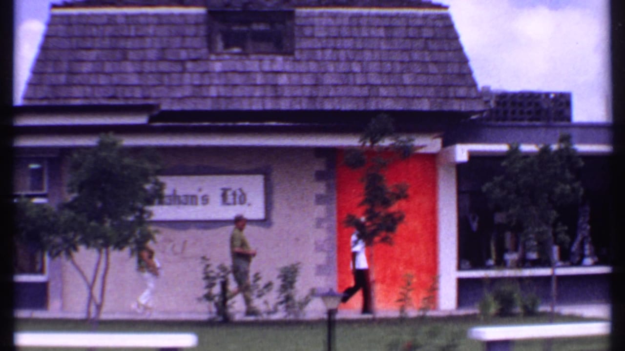 1969 Bahamas home-movie frame showing people walking past a low commercial building with red wall panels, signage, and planted trees.
