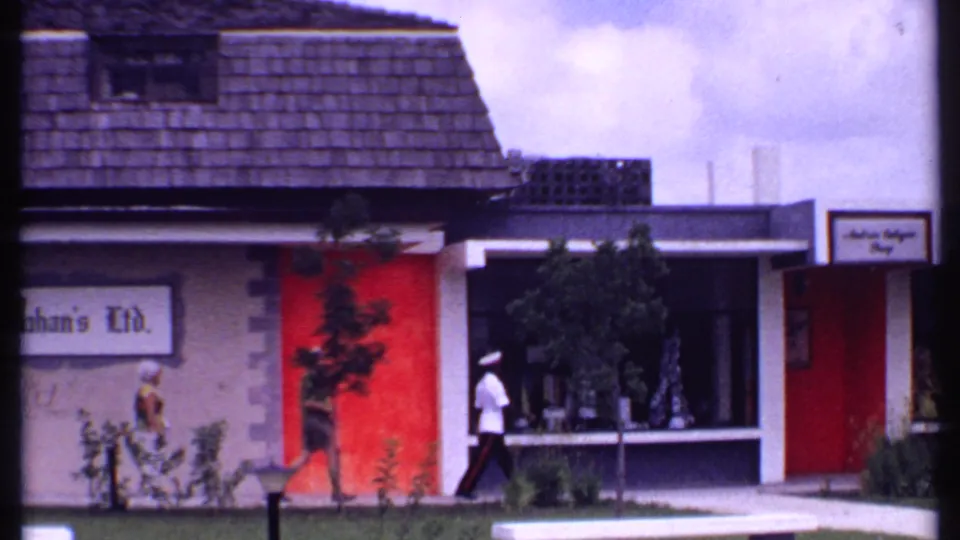 1969 Bahamas street view with a man in white uniform and other pedestrians passing a red-accented storefront and display window.