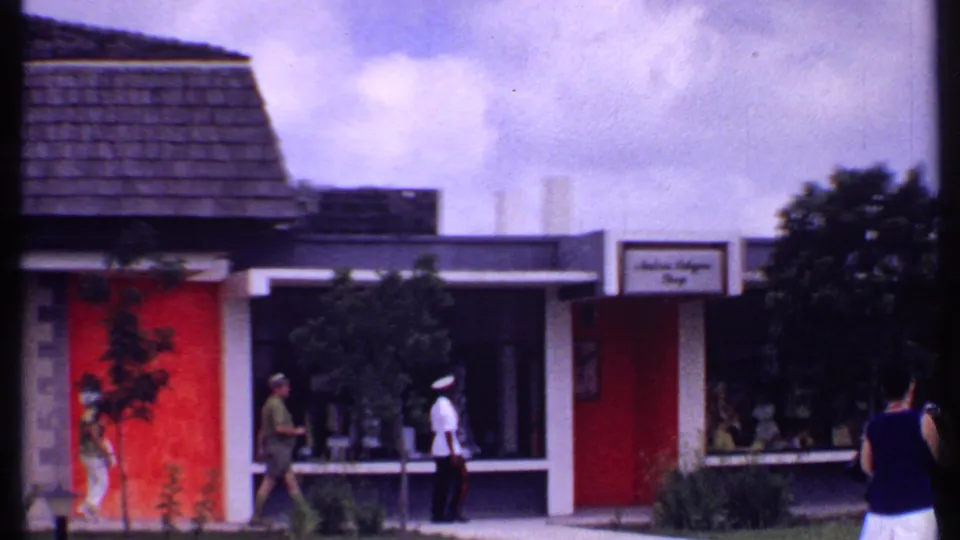 1969 Bahamas home-movie frame showing a woman with a camera near a red-accented storefront and a man in white uniform by the window.