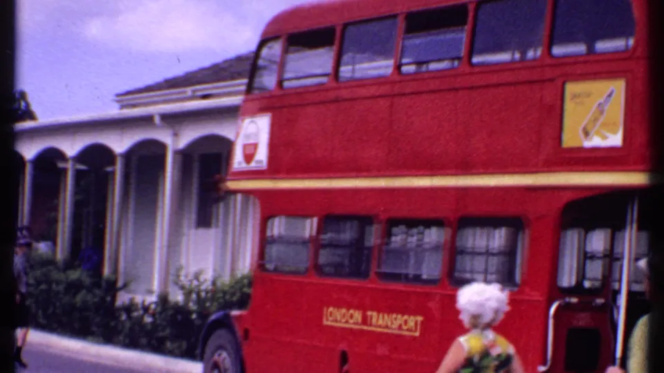 1969 Bahamas street frame with a red London Transport double-decker bus, arched building facade, and a woman holding a camera in the foreground.