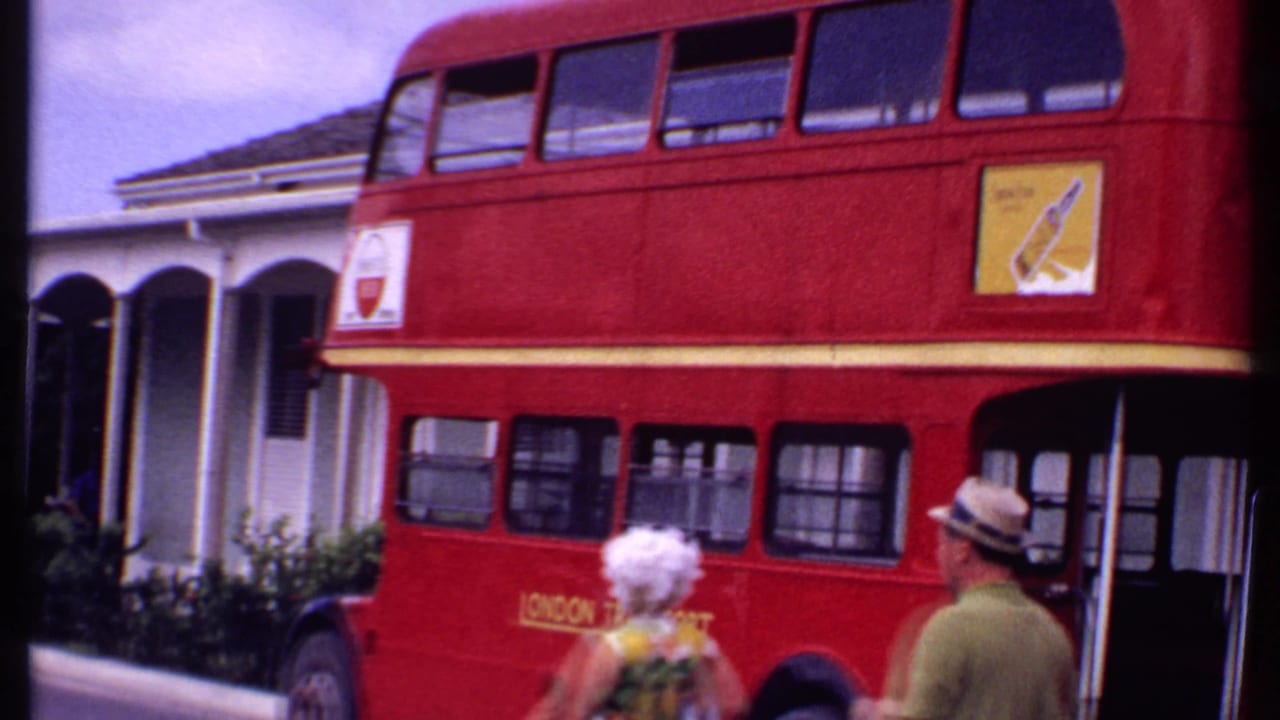 Red London Transport double-decker bus beside a tropical street with two pedestrians in front.