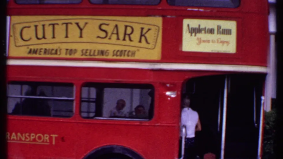 1969 Bahamas frame showing the rear side of the red double-decker bus, alcohol advertisements, passengers, and a person by the open doorway.