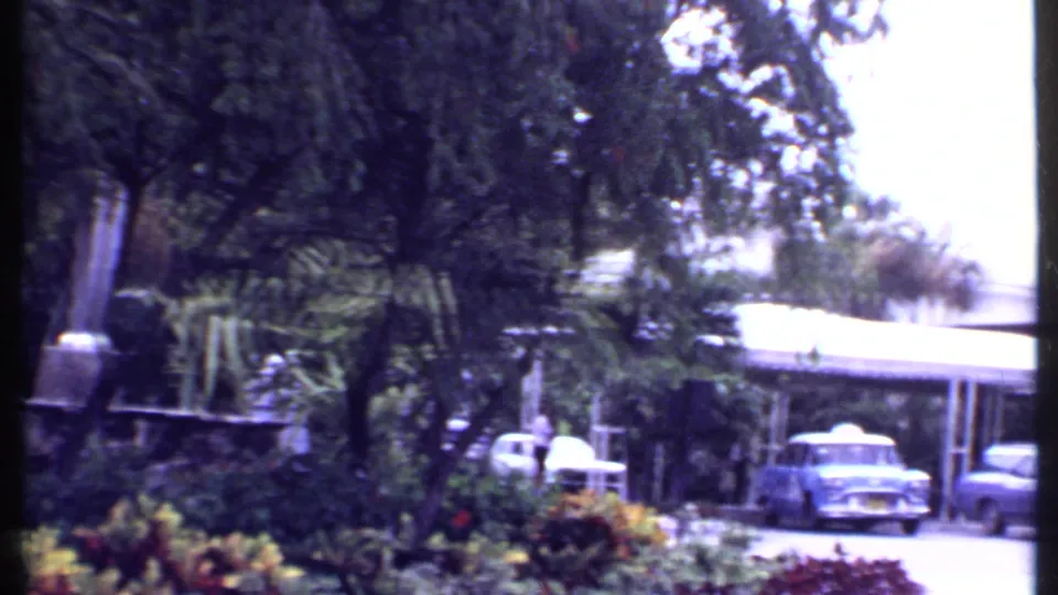 1969 Bahamas home-movie frame showing parked cars under or near a carport beside tropical trees and planted grounds.