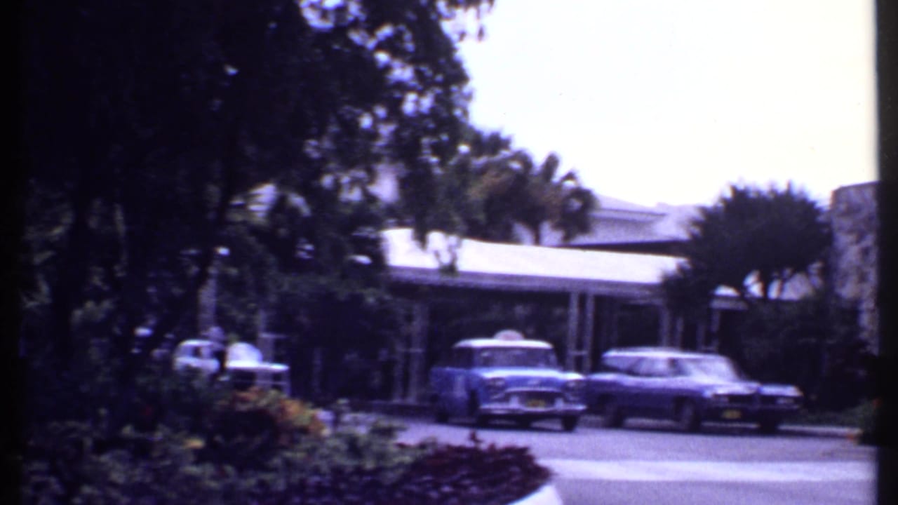 Classic cars outside a tropical resort entrance with stone pillars and palm trees.