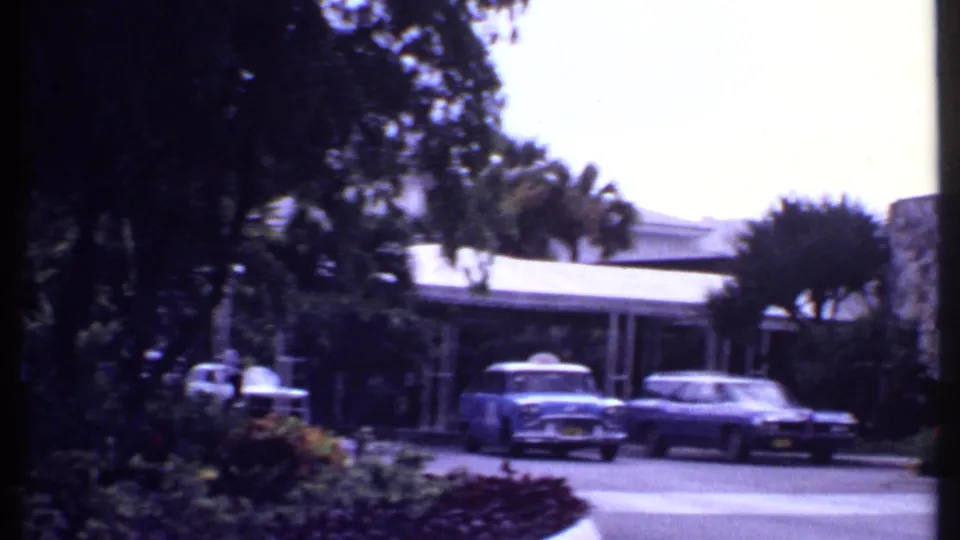 1969 Bahamas home-movie frame showing classic cars near a resort driveway, stone pillars, palms, and landscaped shrubs.