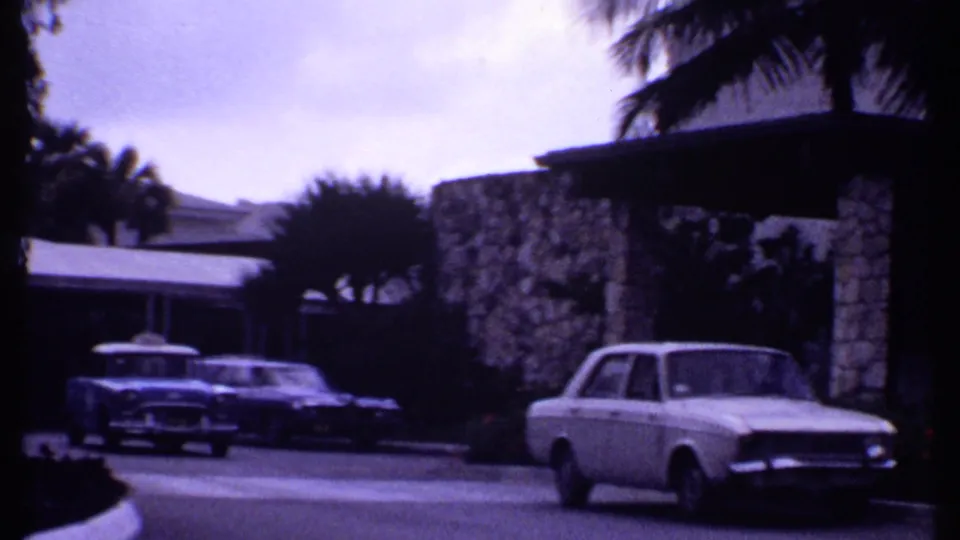 1969 Bahamas frame with a blue taxi and another car near a covered resort entrance framed by stone columns and palms.